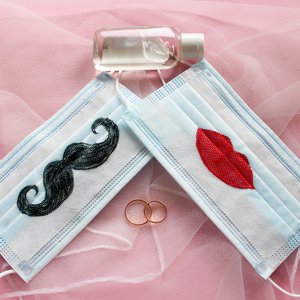 Two wedding rings, antiseptic and protective face masks with a mustache and lips painted on them are on pink background. The concept of wedding ceremonies during a pandemic of coronavirus COVID-19